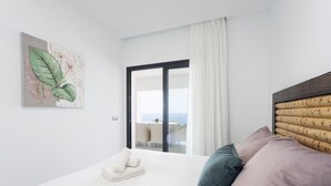 A modern hotel room with a large bed, a view of the sea through a sliding glass door, and a painting of flowers on the wall.
