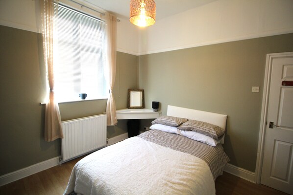 Une chambre à coucher comprenant un lit, une table de chevet, un radiateur, une fenêtre avec des rideaux et une porte.