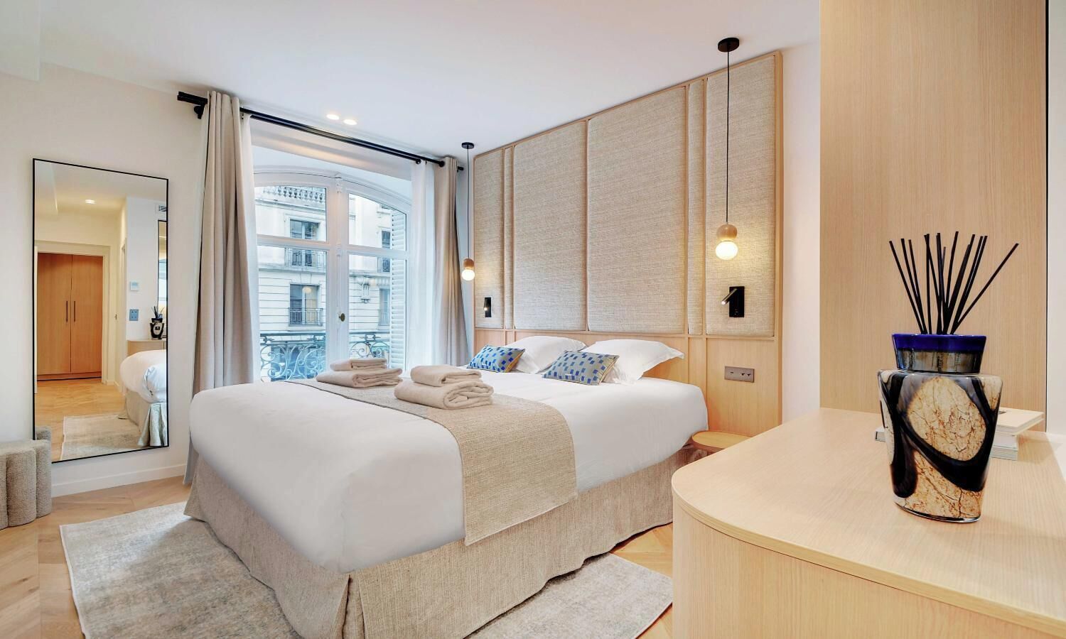 Een moderne hotelkamer met een groot bed, een houten hoofdbord, een nachtkastje, een spiegel en uitzicht op de stad door het raam.