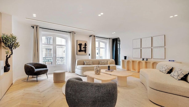 Een moderne woonkamer met een bank, fauteuils en een salontafel. Er zijn grote ramen met gordijnen, een kamerplant en kunst aan de muren.
