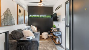 En moderne stue med en grå sofa, et sofabord og et fjernsyn monteret på væggen. Der er en ventilator i loftet, et vindue med udsigt til grønne omgivelser, og en reol med pynteting.