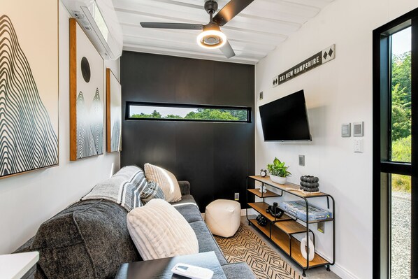 A modern living room with a grey sofa, a large window, a flat-screen TV, and a ceiling fan.