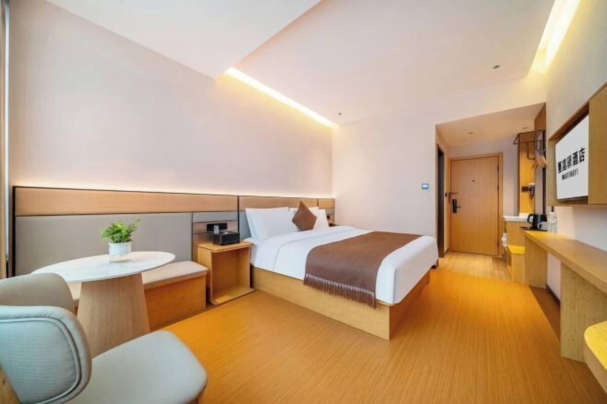 A modern hotel room with a bed, bedside table, chair, and a small round table with a plant.