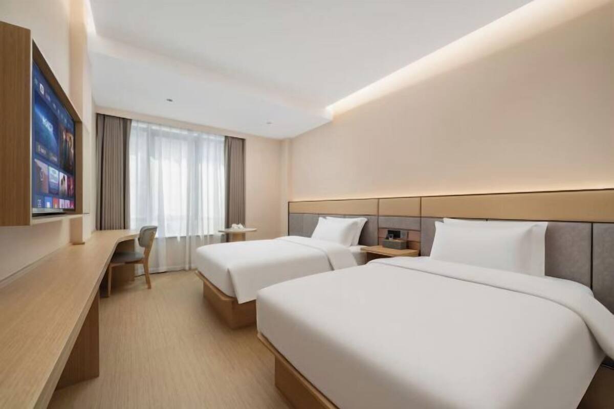 A hotel room with two beds, a desk, a TV, and a large window with curtains.
