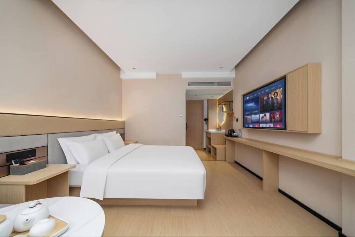 A modern hotel room with a large bed, a TV mounted on the wall, and a small desk with a lamp.