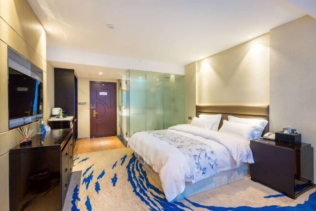 A modern hotel room with a large bed, a TV, a desk, and a glass shower enclosure.