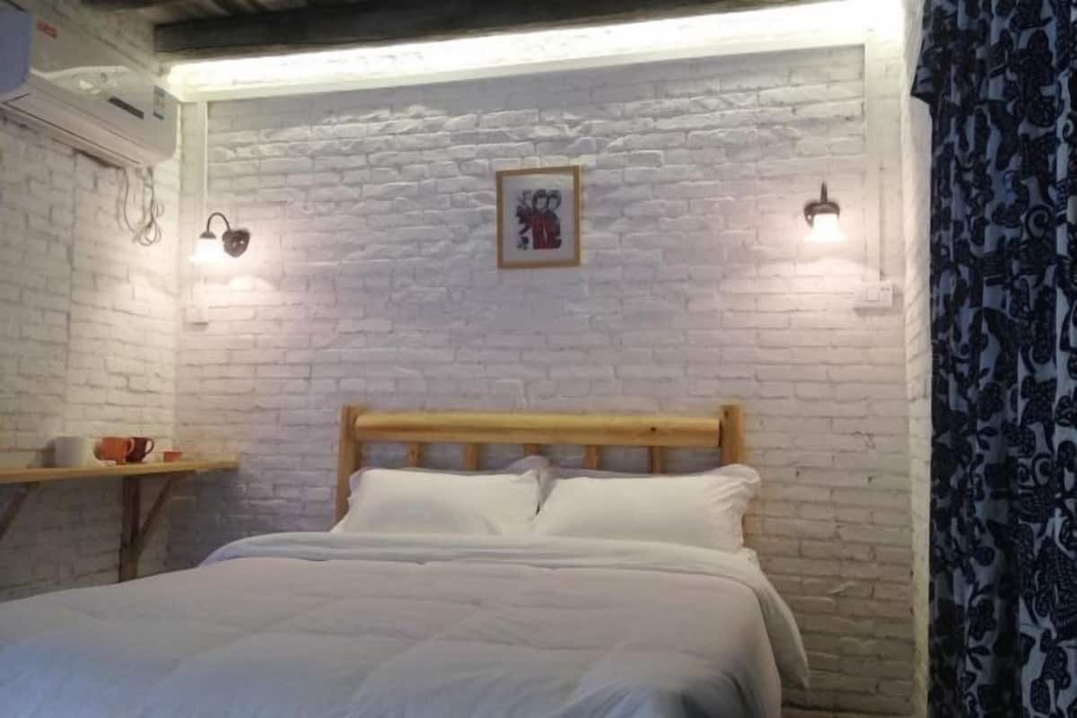 A bedroom with a brick wall, a wooden headboard, a bed with white bedding, two wall-mounted lights, and a small shelf with a cup and a mug.