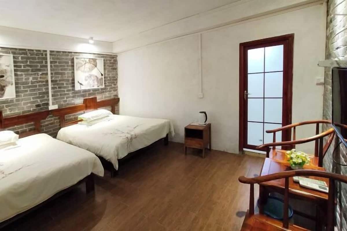 A hotel room with two beds, a wooden table, a small wooden stool, a brick wall with artwork, and a door with frosted glass panels.
