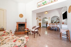Un salon avec une cheminée, un coin repas avec une table et des chaises, et une cuisine visible à travers une arche.