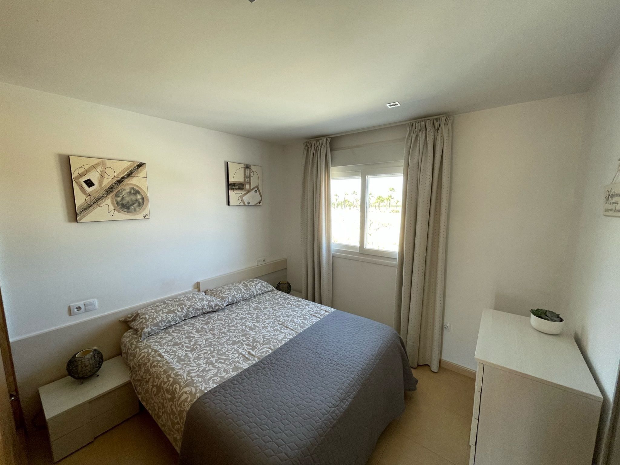 Un dormitorio con cama, mesita de noche, cortinas en la ventana y dos cuadros en la pared.