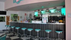 Een bar met een neonbord, barstoelen en een toonbank vol flessen en glazen.