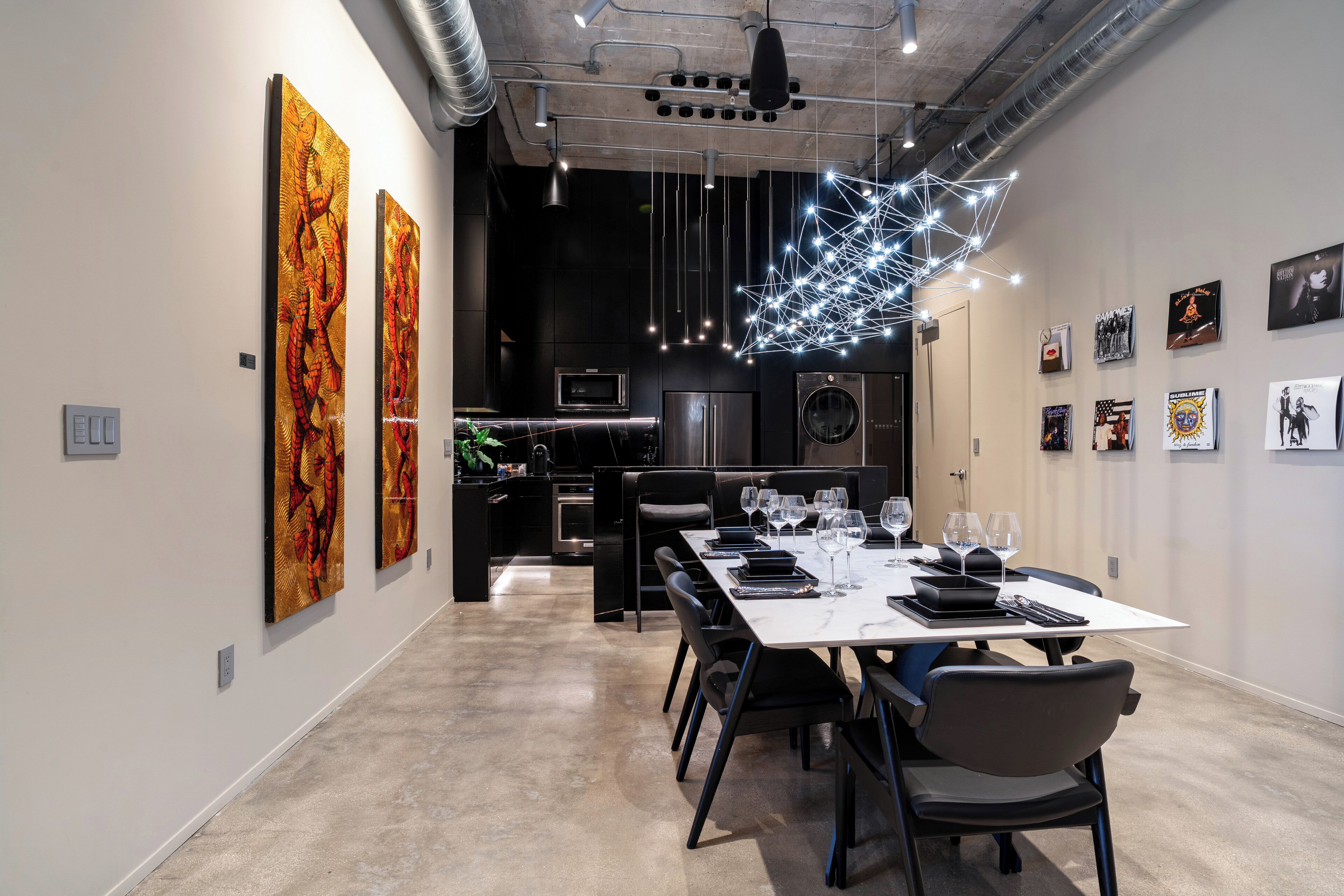 A modern dining area with a long table set for six, a kitchen in the background, and artwork on the walls.