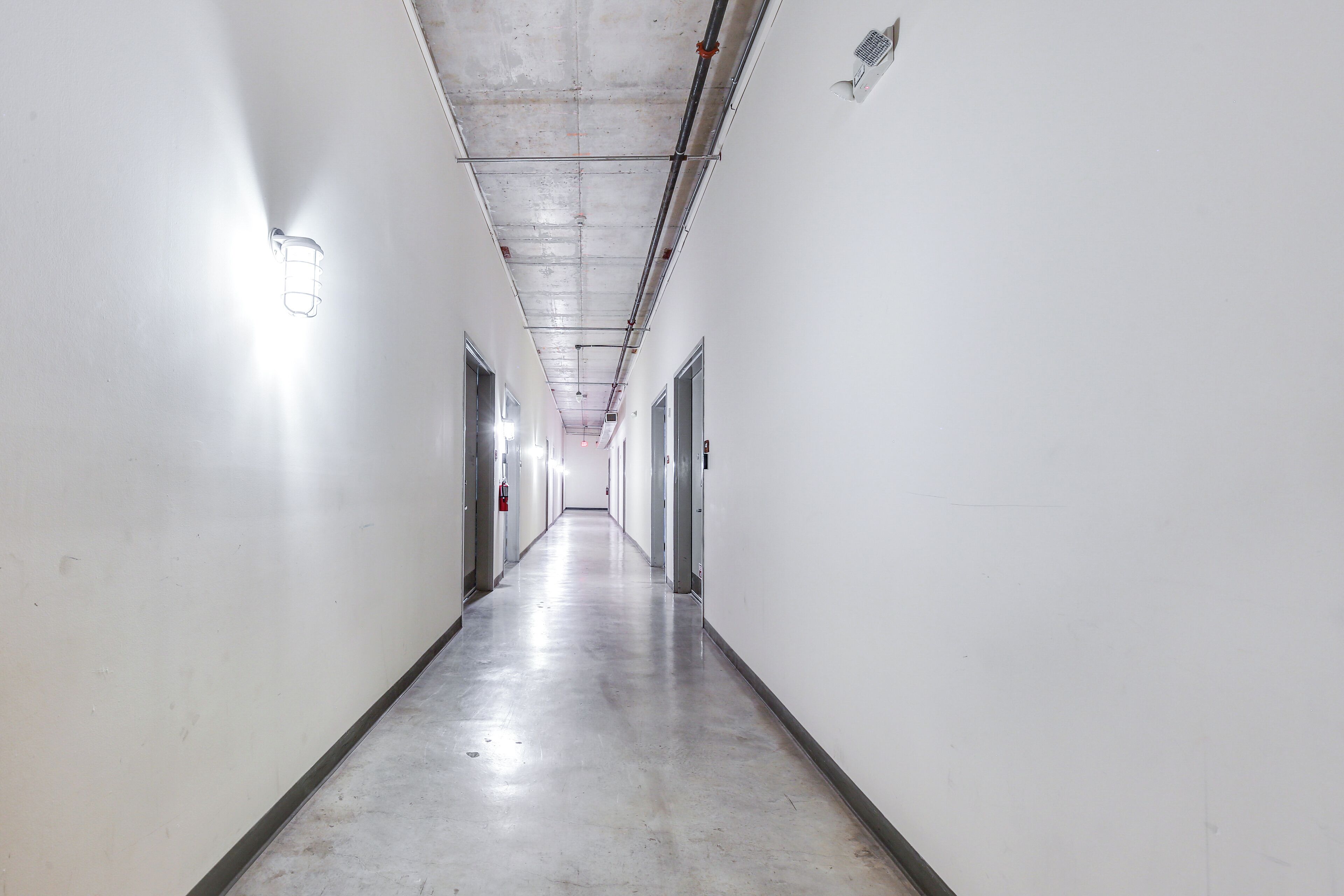 A long, narrow hallway with white walls and a concrete floor, featuring doors and a fire extinguisher on the left.