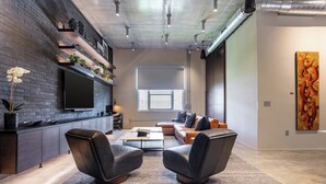 A modern living room with a brick wall, a flat-screen TV, leather chairs, a coffee table, and a large window. - Wynwood Condo, Walk to Restaurants & Nightclubs! (Miami)