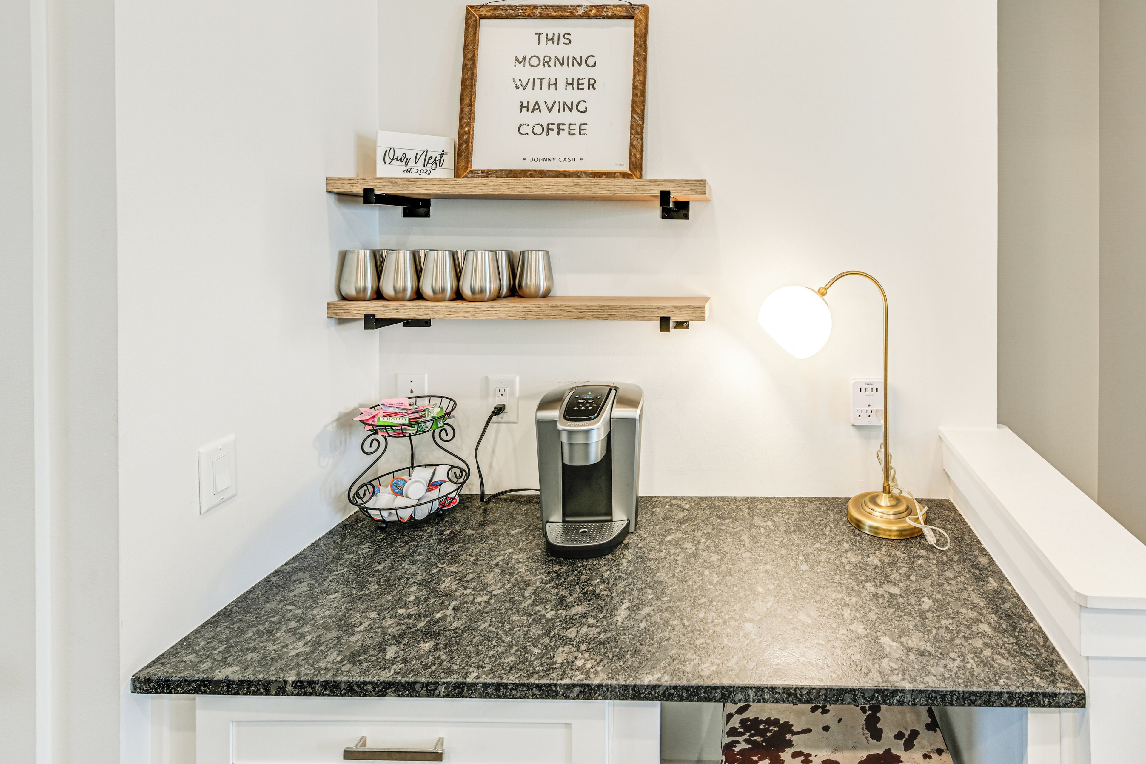 Une cuisine dotée d’un plan de travail en granit, d’une machine à café, d’une lampe et d’étagères où sont posées des tasses et un cadre contenant une citation.