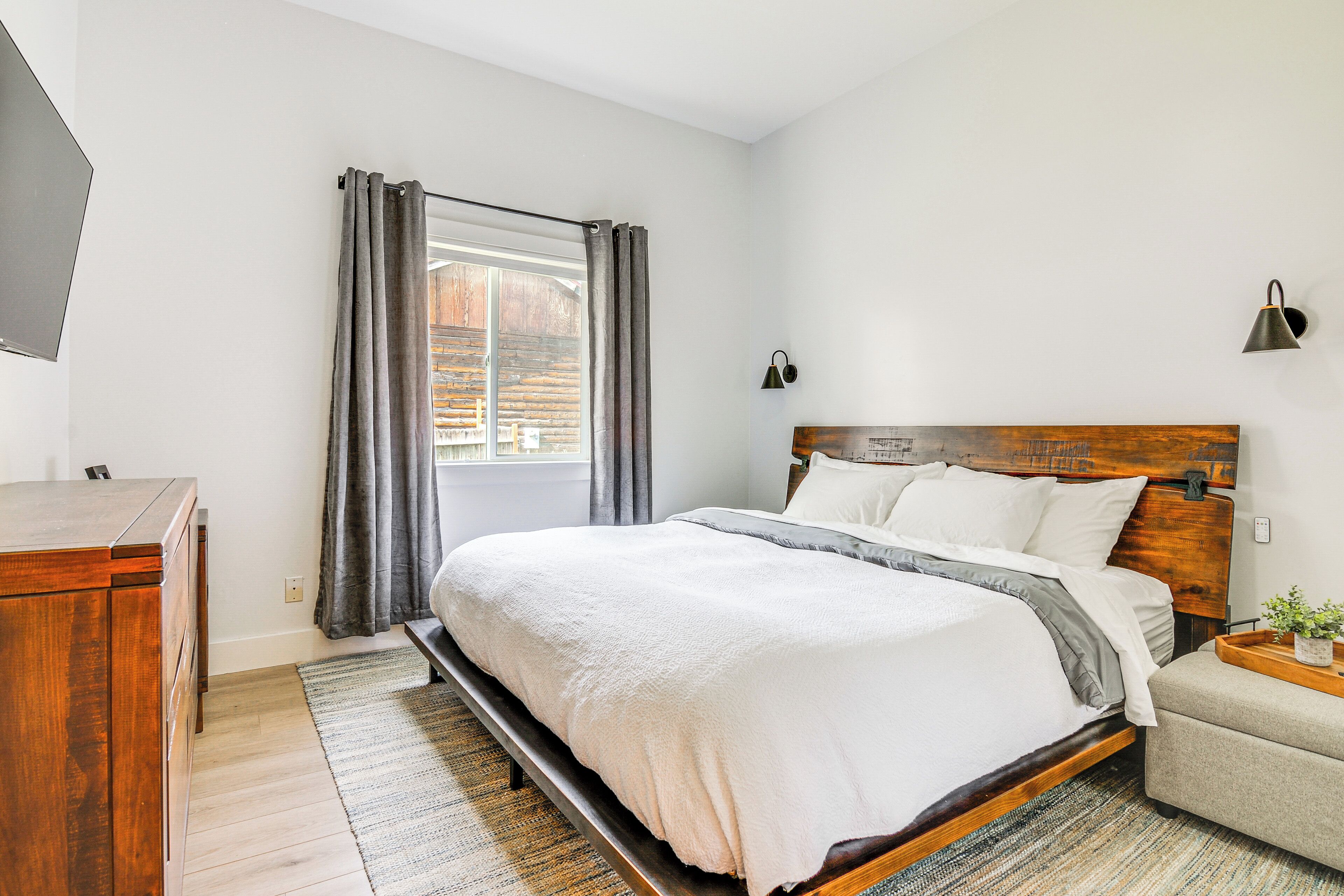 A bedroom with a wooden bed, a wooden dresser, a nightstand, a lamp, and a window with curtains.