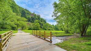 A wooden bridge over a concrete path leading to a grassy area with trees and a hill in the background. - 24 Private Acres: Trails/hot Tub Near Blowing Rock (Lenoir)