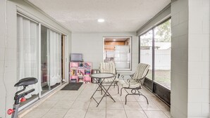 Ein überdachter Patio-Bereich mit einer Schiebetür, einem kleinen Tisch mit Stühlen, einem feststehenden Ergometer und einer Küchenzeile mit Schränken und einem Kühlschrank.