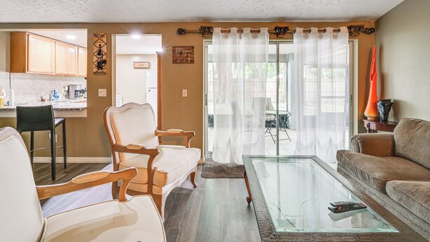 Ein Wohnzimmer mit einem Glastisch, zwei weißen Sesseln, einem braunen Sofa und einer Küche im Hintergrund.