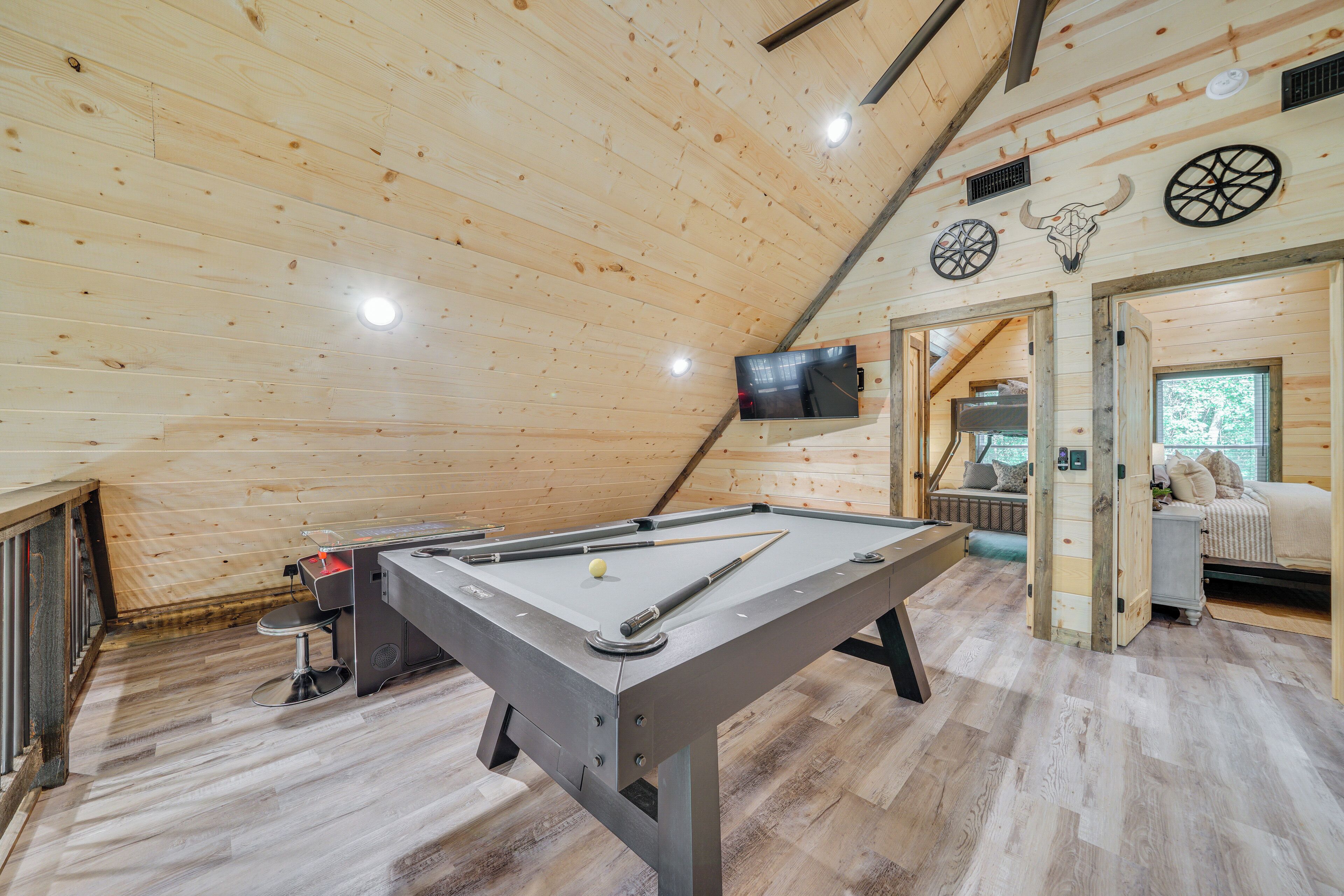 A wooden-floored room with a pool table, a TV mounted on the wall, and a bed in the background.