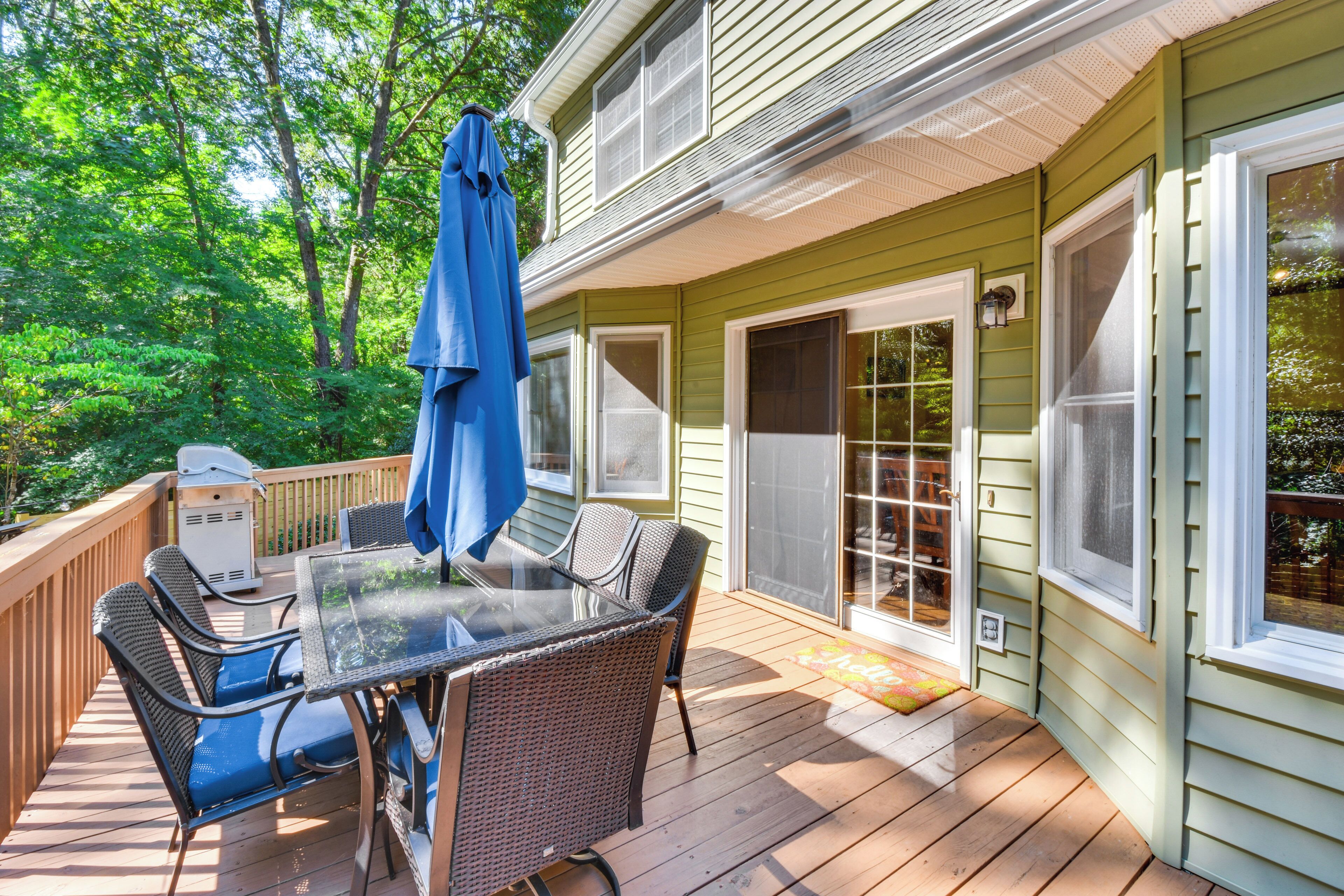 A covered deck with a glass-top table and wicker chairs, surrounded by a wooden deck and a green house.