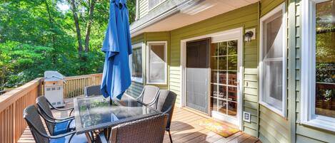 A covered deck with a glass-top table and wicker chairs, surrounded by a wooden deck and a green house.