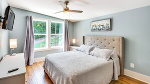 A bedroom with a bed, a ceiling fan, a window with curtains, a bedside table with a lamp, and a painting on the wall. - Waterfront Hartwell Apartment w/ Dock Access! (Hartwell)