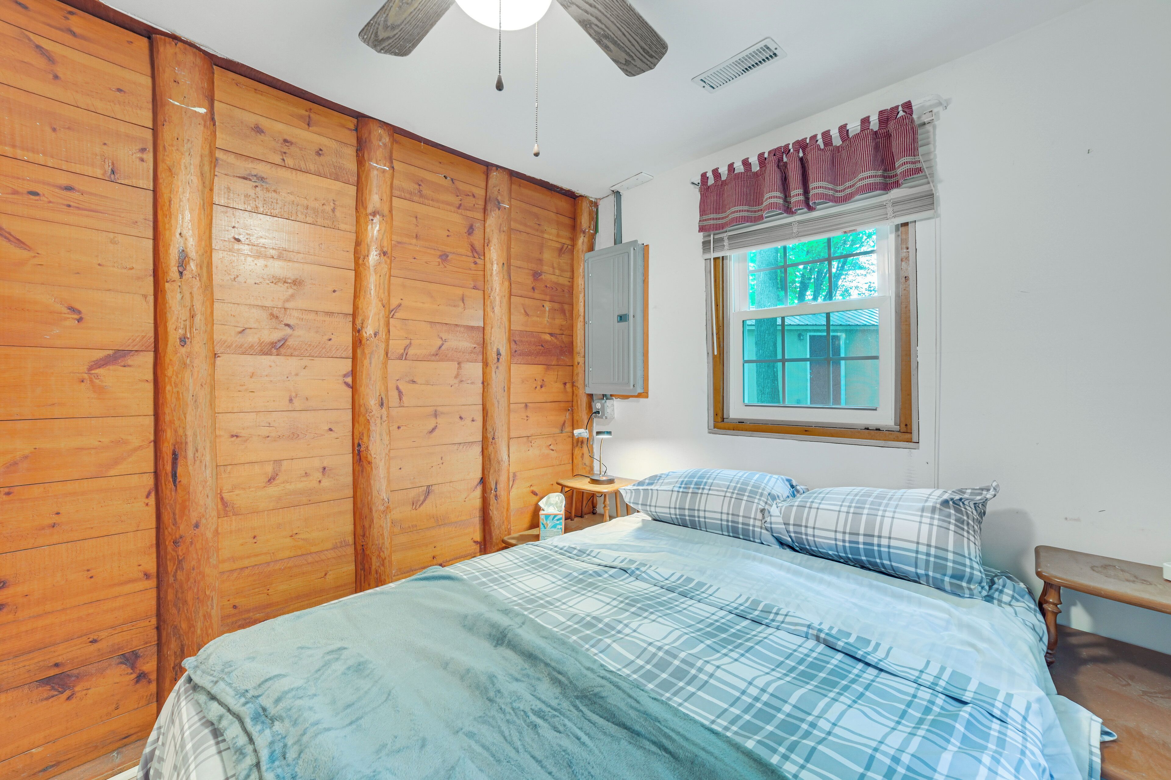 Un dormitorio con paneles de madera, una cama con ropa de cama azul y blanca, una ventana con vistas y un ventilador de techo.