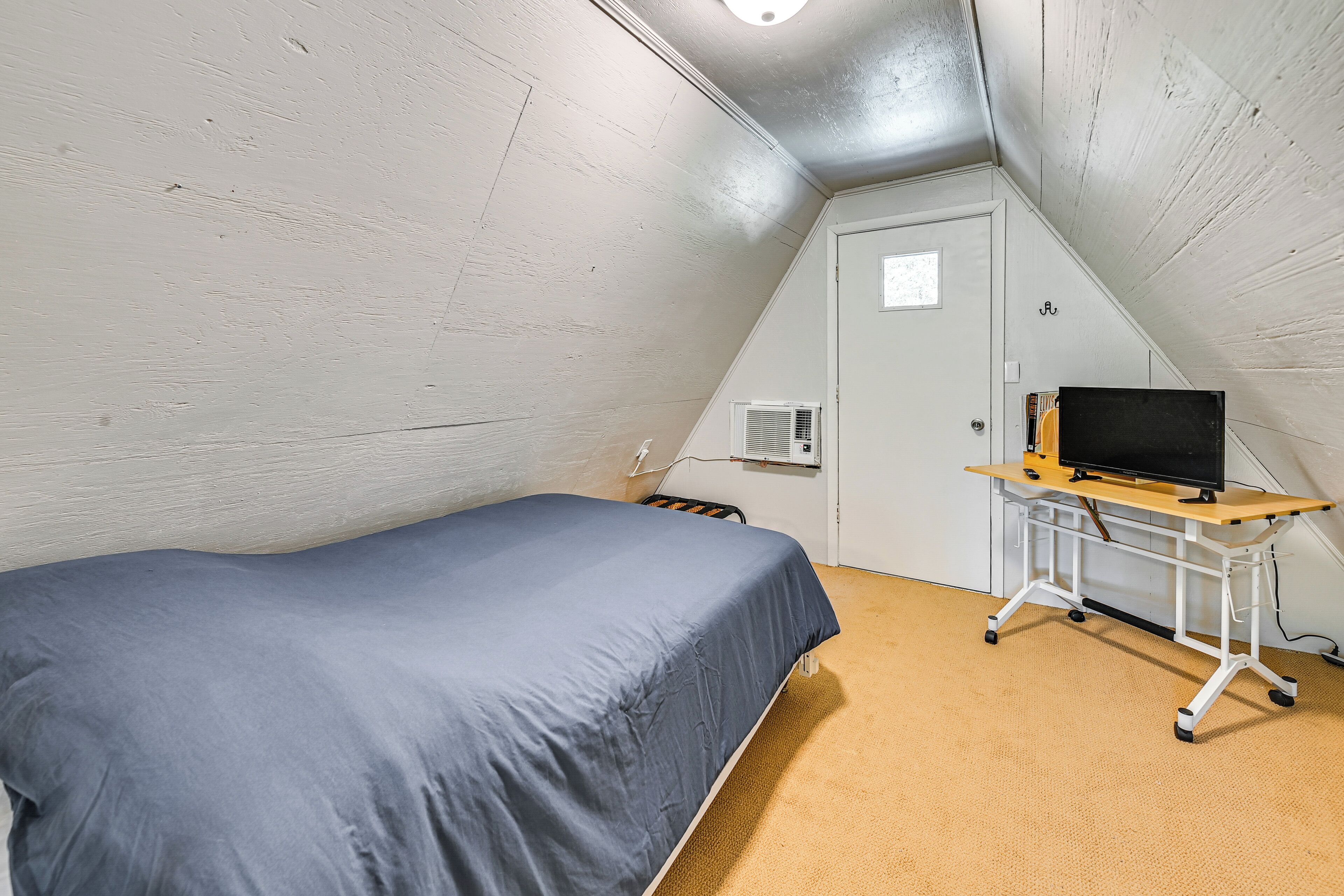 Ein kleiner, dreieckiger Raum mit einem Einzelbett, einem Schreibtisch mit Computer und einer Tür.
