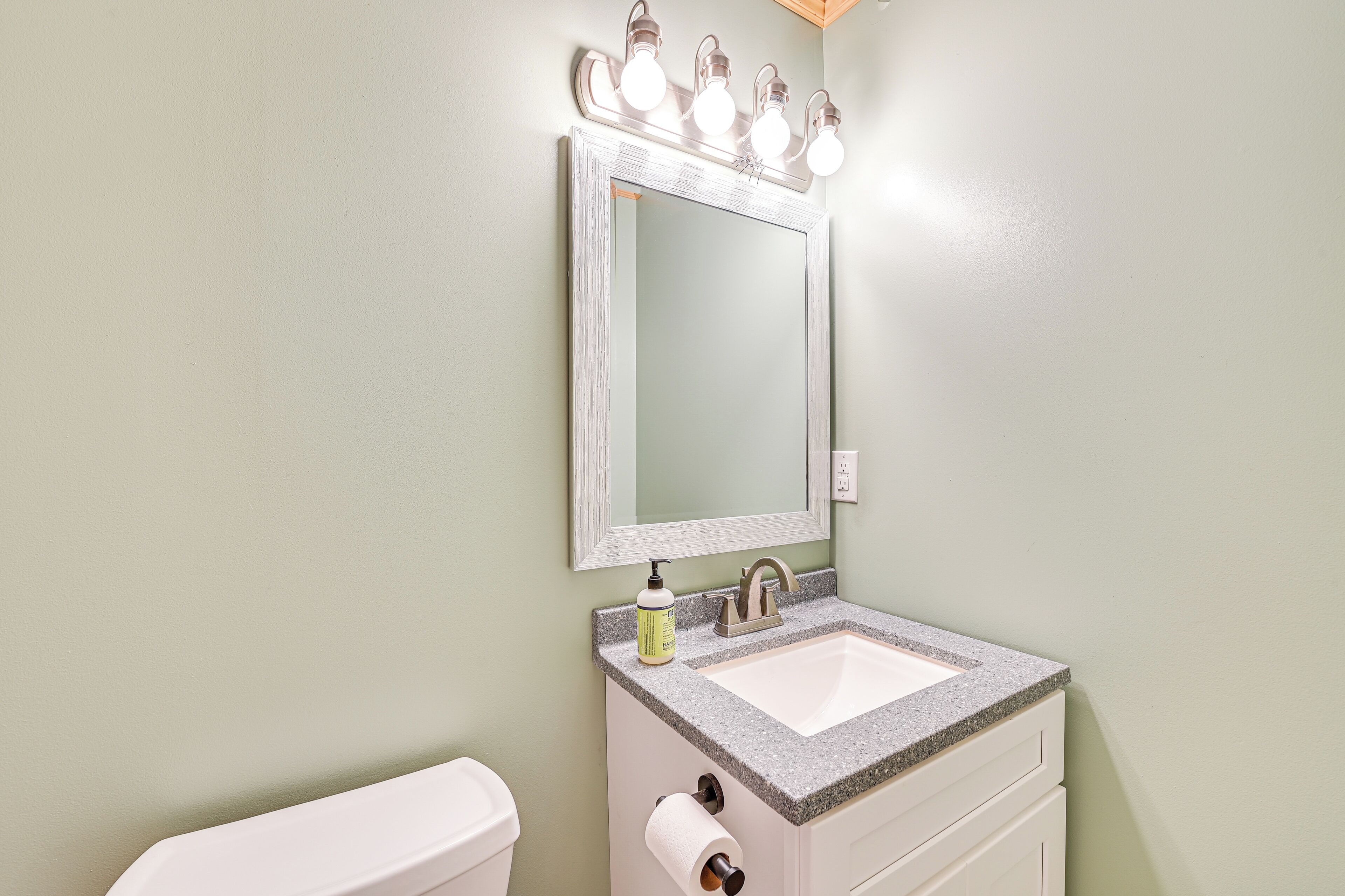A bathroom with a white sink, a mirror with lights, a toilet, and a soap dispenser.
