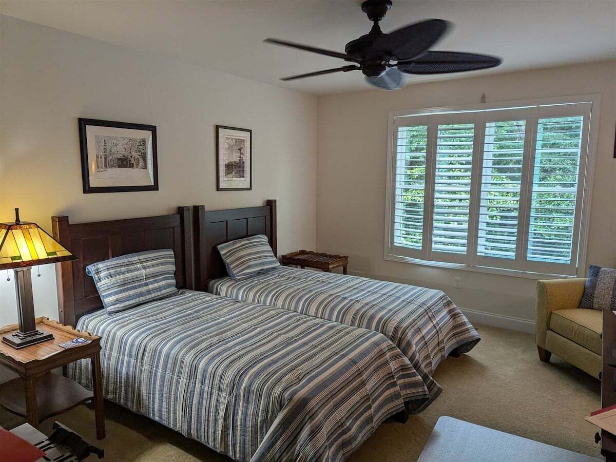 A bedroom with two beds, a ceiling fan, a window with blinds, and a lamp on a bedside table.