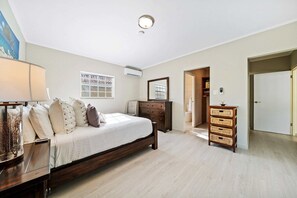 A bedroom with a bed, dresser, and a mirror. There is a window with a view, an air conditioning unit, and a lamp.