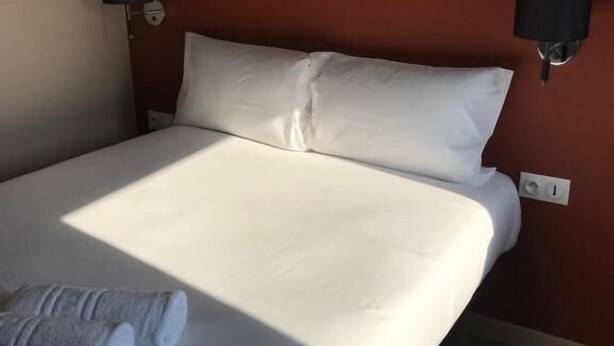 A neatly made bed with white linens and two pillows, a dark brown wall, and two wall-mounted lamps on either side of the bed. - Appart'Hotel Voltaire  (Chateauroux)