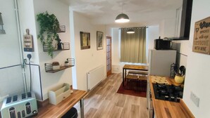 A compact kitchen with a dining area, wooden flooring, and a window with curtains. - Bramble Cottage Rhondda Valley by STAE-Homes (Tylorstown)