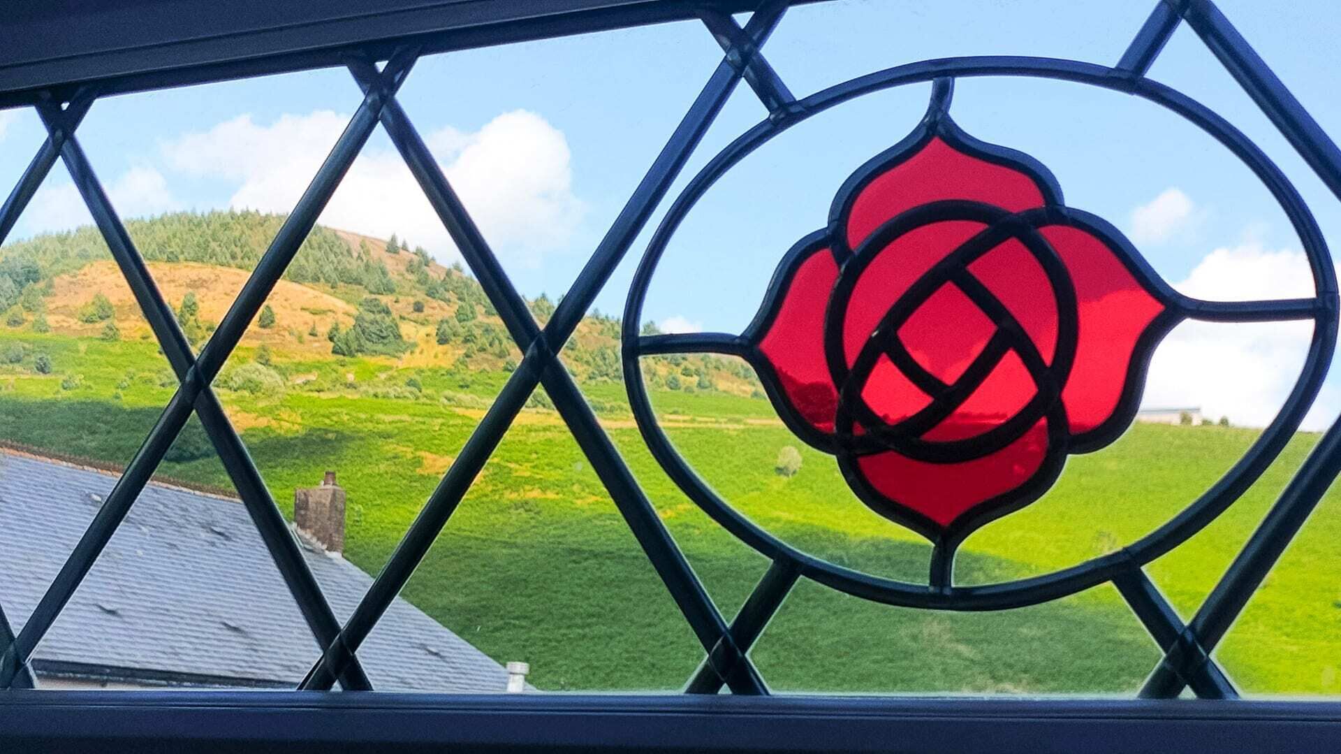 A window with a geometric pattern and a red rose design, overlooking a green landscape with hills.