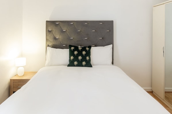 A bed with a tufted headboard, white bedding, a green pillow with a floral pattern, and a bedside lamp.