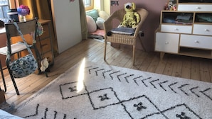 A cozy room with a wooden floor, a white rug with geometric patterns, a wicker chair, a white dresser, and a wall with alphabet posters and photos.