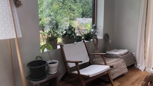 A room with a wooden floor, a white woven chair, a small table with potted plants, and a view of greenery outside.
