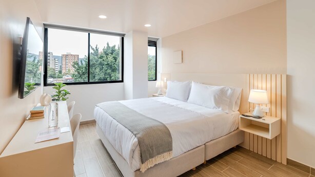 A modern hotel room with a large bed, bedside tables, a desk with a chair, a TV, and a window with a view of buildings and trees.