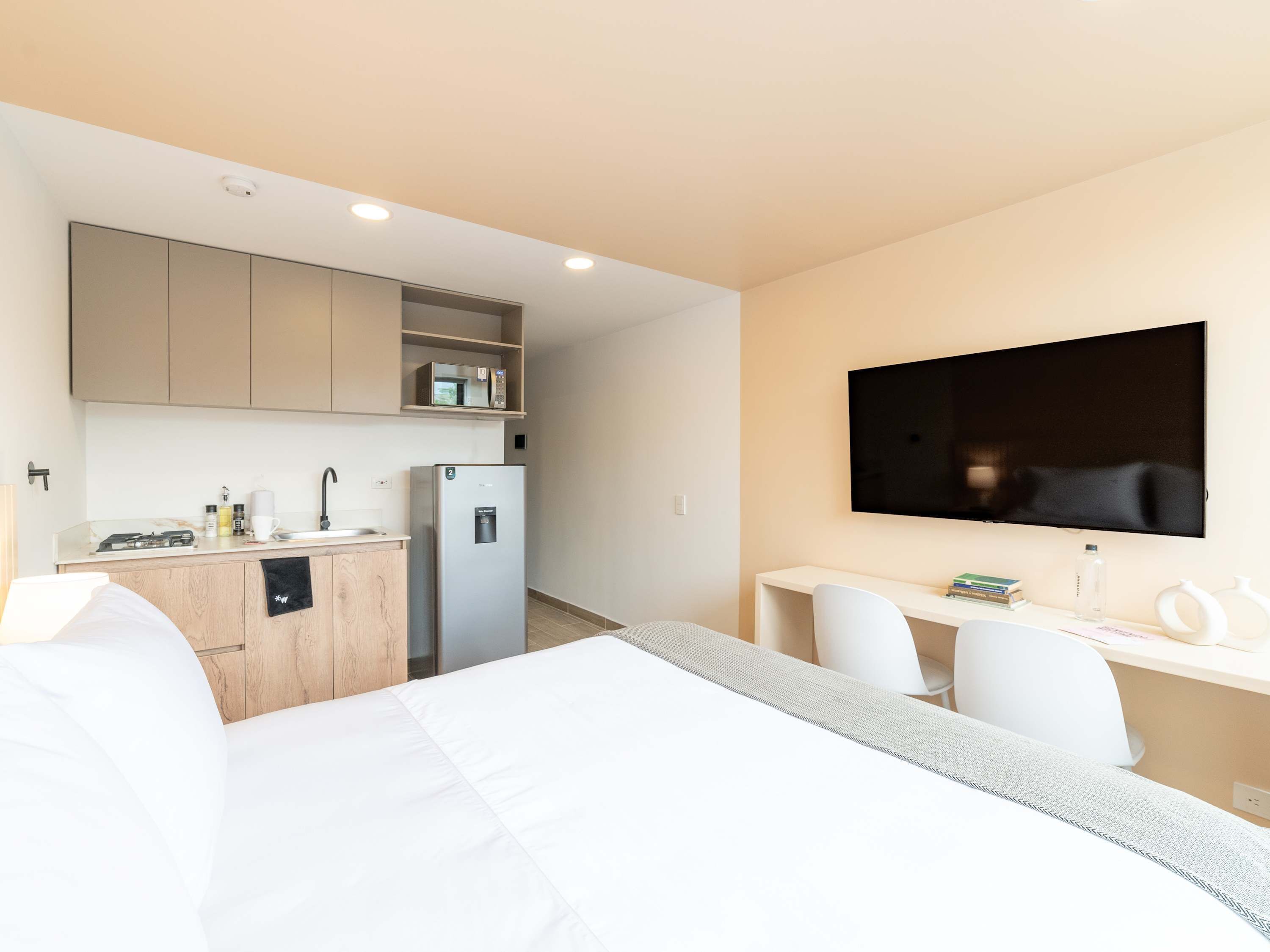 A modern hotel room with a bed, a desk, a flat-screen TV, a refrigerator, and a kitchenette.