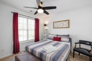 Une chambre à coucher avec un lit, un ventilateur de plafond, une fenêtre avec des rideaux rouges, une table de chevet avec une lampe et un tableau encadré au mur.