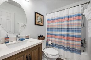Une salle de bain avec un lavabo blanc, un miroir et des toilettes. On y trouve un rideau de douche à rayures et un tableau encadré accroché au mur.