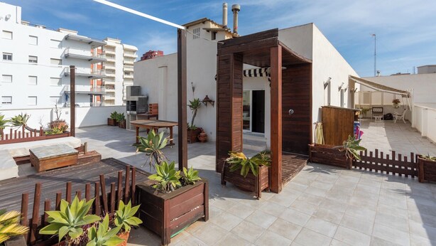 Una terraza en la azotea con una pérgola de madera, plantas en macetas, un espacio de estar y vistas a edificios cercanos.