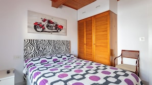 Un dormitorio con cabecera de tablero de ajedrez, cama con colcha de motivos geométricos, armario de madera y un cuadro de una motocicleta en la pared.