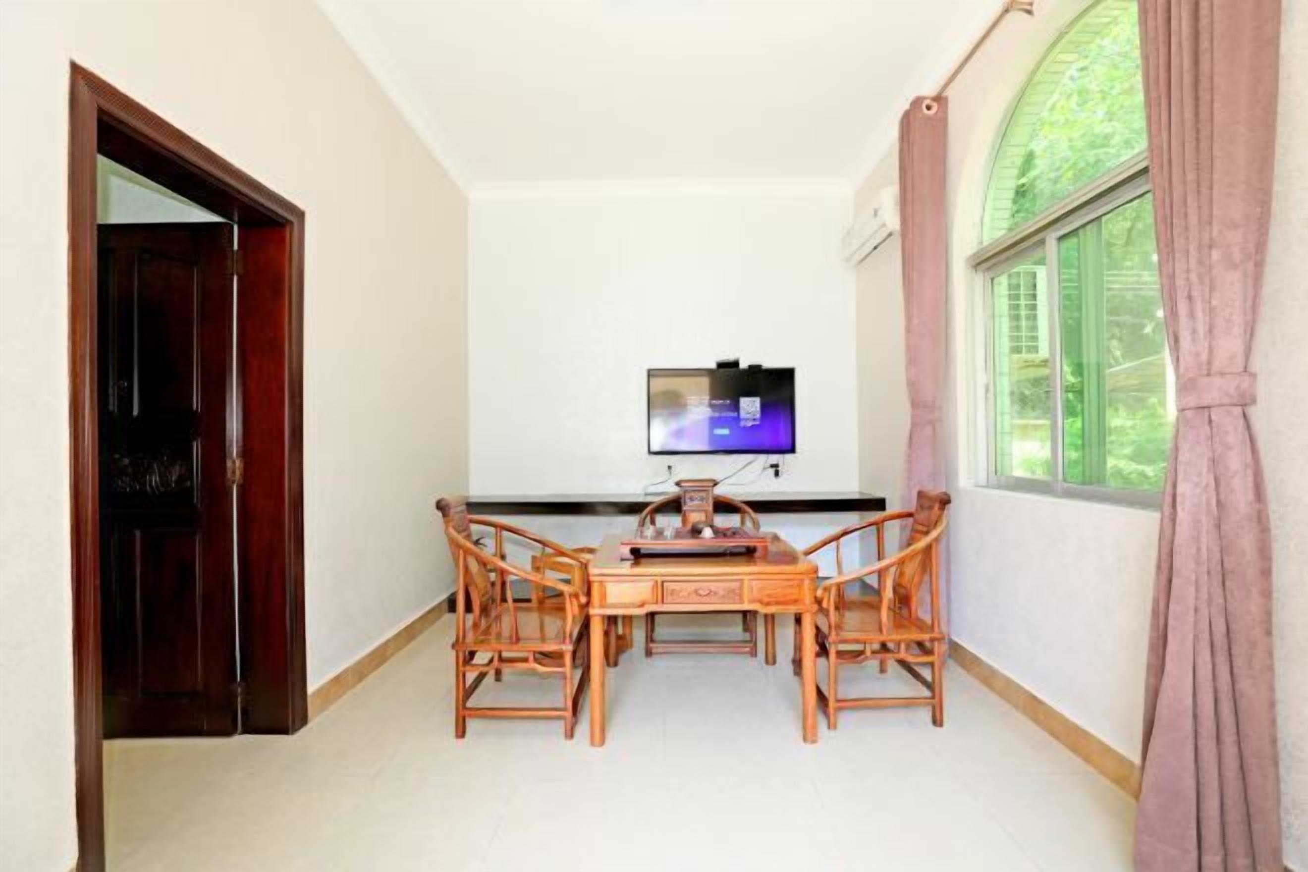 A room with a wooden dining table and chairs, a television on the wall, and a window with curtains.
