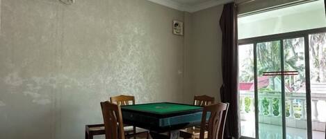 A room with a pool table, four wooden chairs, a ceiling fan, and a large window with a view of greenery.