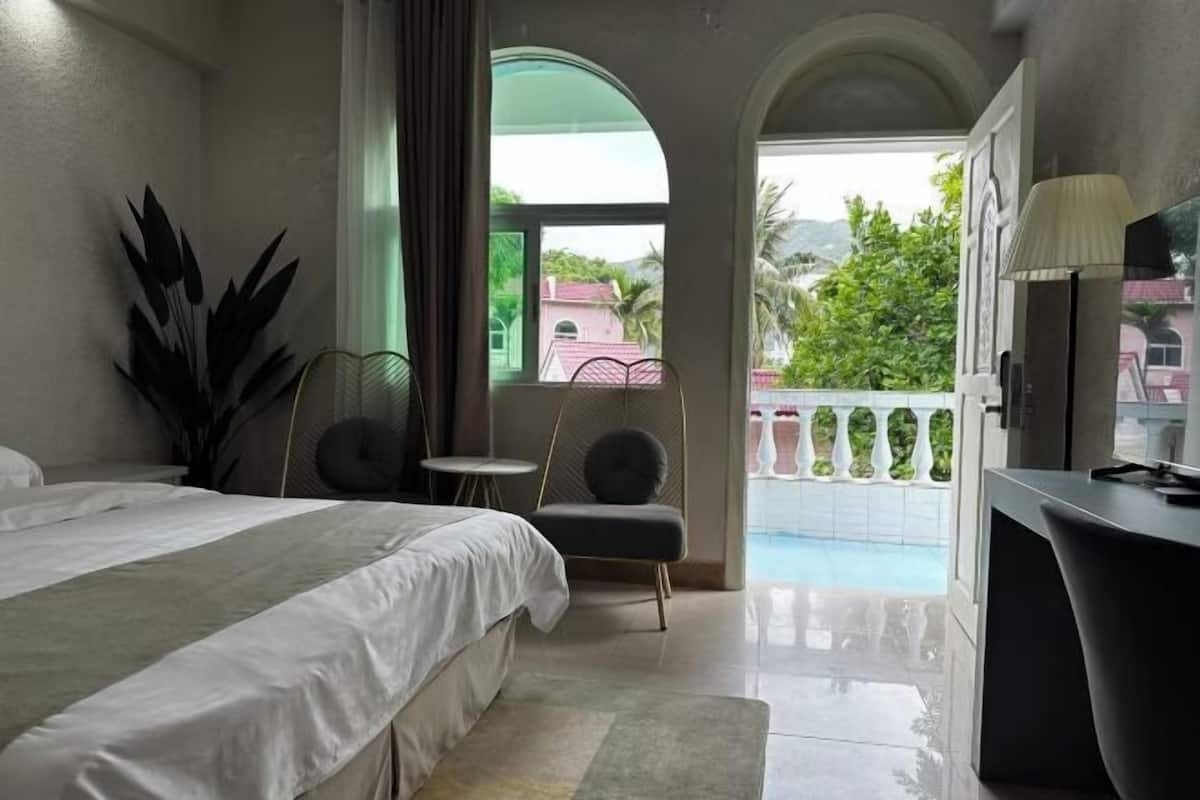 Une chambre d’hôtel comprenant un lit, un bureau, une chaise, une plante en pot et un balcon donnant sur un paysage verdoyant et des bâtiments.