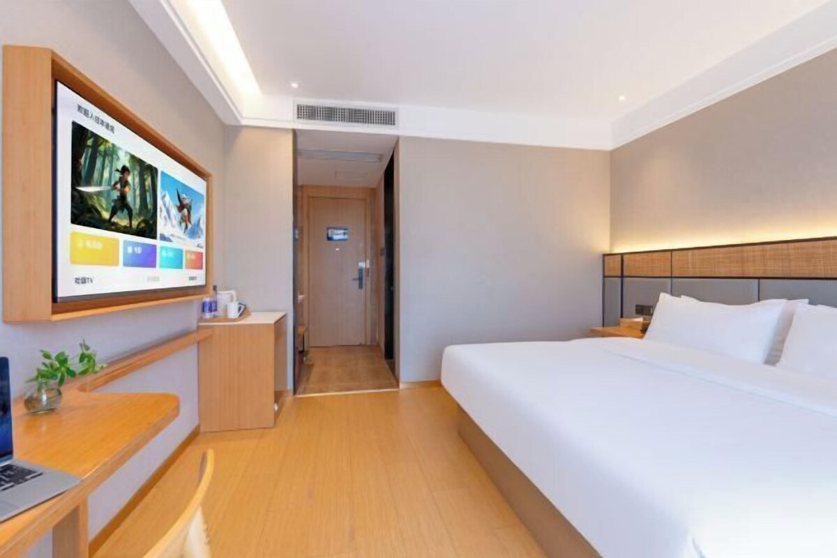 A hotel room with a bed, a desk with a laptop, a TV on the wall, and a door leading to another room.