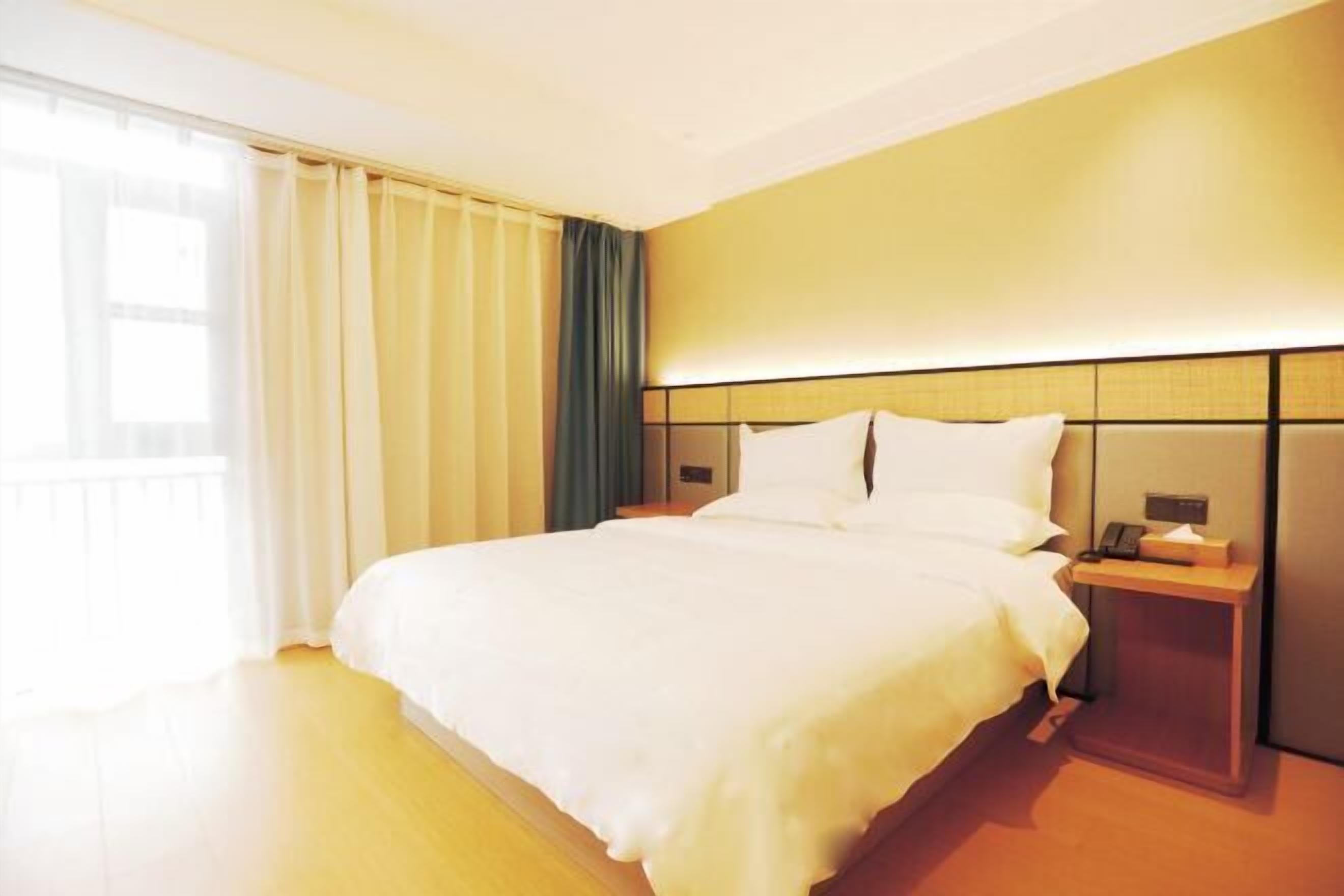 Habitación de hotel con una cama grande, una mesita de noche con lámpara y vistas al exterior a través de una ventana con cortinas.