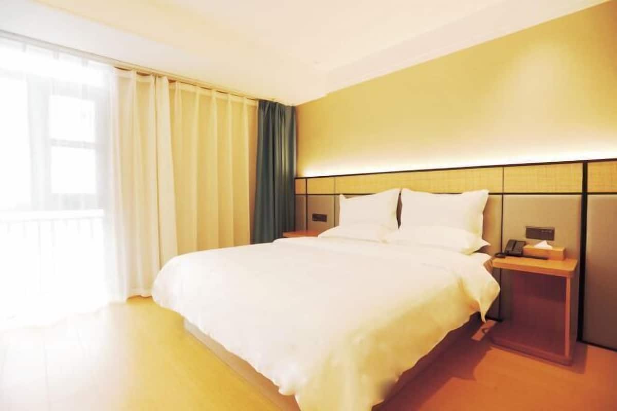 Habitación de hotel con una cama grande, una mesita de noche con lámpara y vistas al exterior a través de una ventana con cortinas.